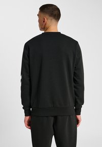 Man med kort hår som bär en enkel svart sweatshirt och svarta byxor, står vänd bort från kameran mot en enkel ljus bakgrund.