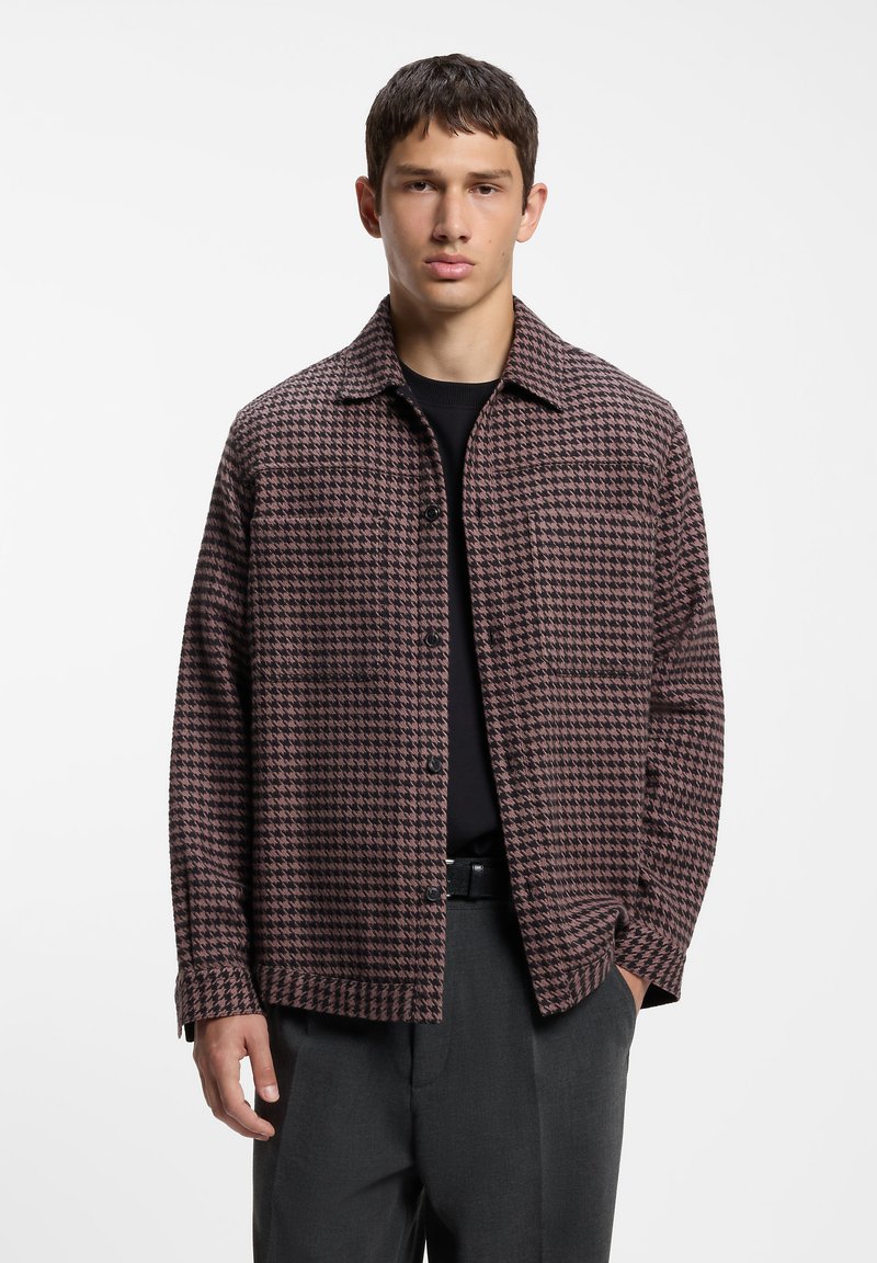 Jeune homme portant une veste boutonnée à motif pied-de-poule marron et noir, une chemise noire et un pantalon gris foncé sur fond blanc.