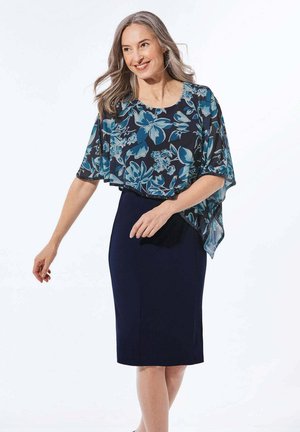 Lächelnde Frau mit grauen Haaren trägt einen marineblauen Bleistiftrock und ein marineblaues, blaues und türkisfarbenes florales transparentes Cape-Oberteil und posiert vor weißem Hintergrund.