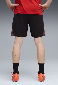 Czarne, sportowe szorty z teksturowanym bocznym panelem z pomarańczowymi i czerwonymi wzorami. Noszone z pomarańczowymi korkami i czarnymi skarpetkami.