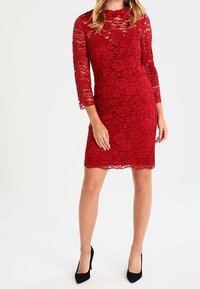 Vestito rosso in pizzo con maniche lunghe, silhouette aderente, motivi floreali, scollatura a girocollo e lunghezza sopra il ginocchio. Abbinato a tacchi alti neri.