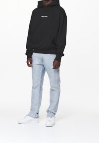 Svart hoodie med framficka och vit text "PEGADOR", kombinerad med ljusblå jeans och vita sneakers, stående mot en enkel bakgrund.