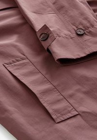 Manche de veste mauve pliée avec un bouton marron et gros plan sur une poche rectangulaire cousue sur le tissu mauve.