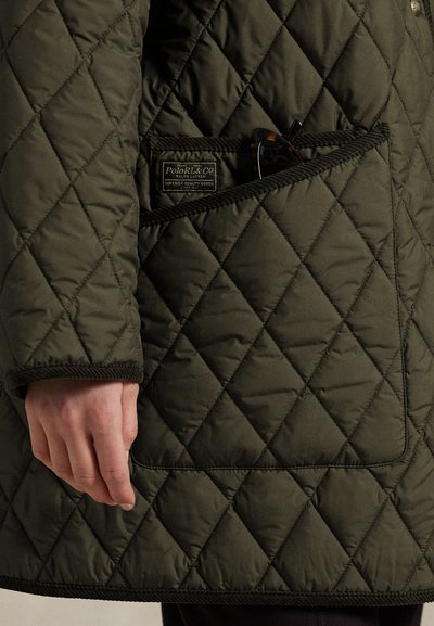 Õmblustega oliivrohelise jope taskus on kilpkonnakujulised päikeseprillid ja Polo Ralph Laureni silt, näha on käsi tasku lähedal.