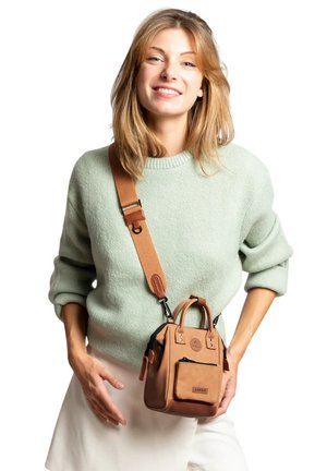 Junge Frau trägt einen hellgrünen Pullover und weiße Hose, hält eine kleine braune Umhängetasche mit breitem Riemen und lächelt.