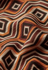 Gros plan d’un tissu avec des motifs géométriques carrés concentriques marron, orange, noir et blanc, montrant la texture et les plis.