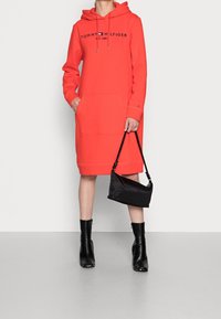 Korallfärgad sweatshirtklänning med huva, ficka fram och logotyp. Bärs med svarta ankelskor och en liten svart handväska. Slät textur och avslappnad design.