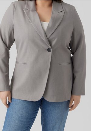 Kvinde iført en lysegrå blazer med en enkelt knap over en hvid top og blå jeans mod en ensfarvet baggrund.