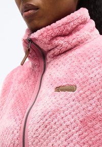 Άτομο που φοράει ροζ υφασμάτινο μπουφάν φλίς με υψηλό γιακά και φερμουάρ, με ένα καφέ λογότυπο Columbia Sportswear στο στήθος.