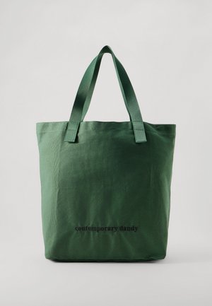 Sac fourre-tout en toile vert foncé, doté de longues anses et portant les mots « contemporary dandy » imprimés sur le devant.