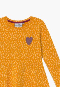 Keltaiset pitkät hihat mekko, jossa on valkoinen kukkakuosi ja kimaltava purppuranvärinen sydänpatsas edessä. Puuvillamateriaali, ympyräkauluksella.