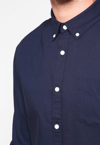 Mörkblå skjorta med knappar i bomull, med krage, vita knappar och en bröstficka med texturerad tyg.