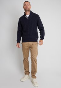 Granatowy sweter z dzianiny z kołnierzem szalowym i przyciskami, zestawiony z jasnymi khaki spodniami i białymi trampkami. Gładka tekstura, dopasowany fason.