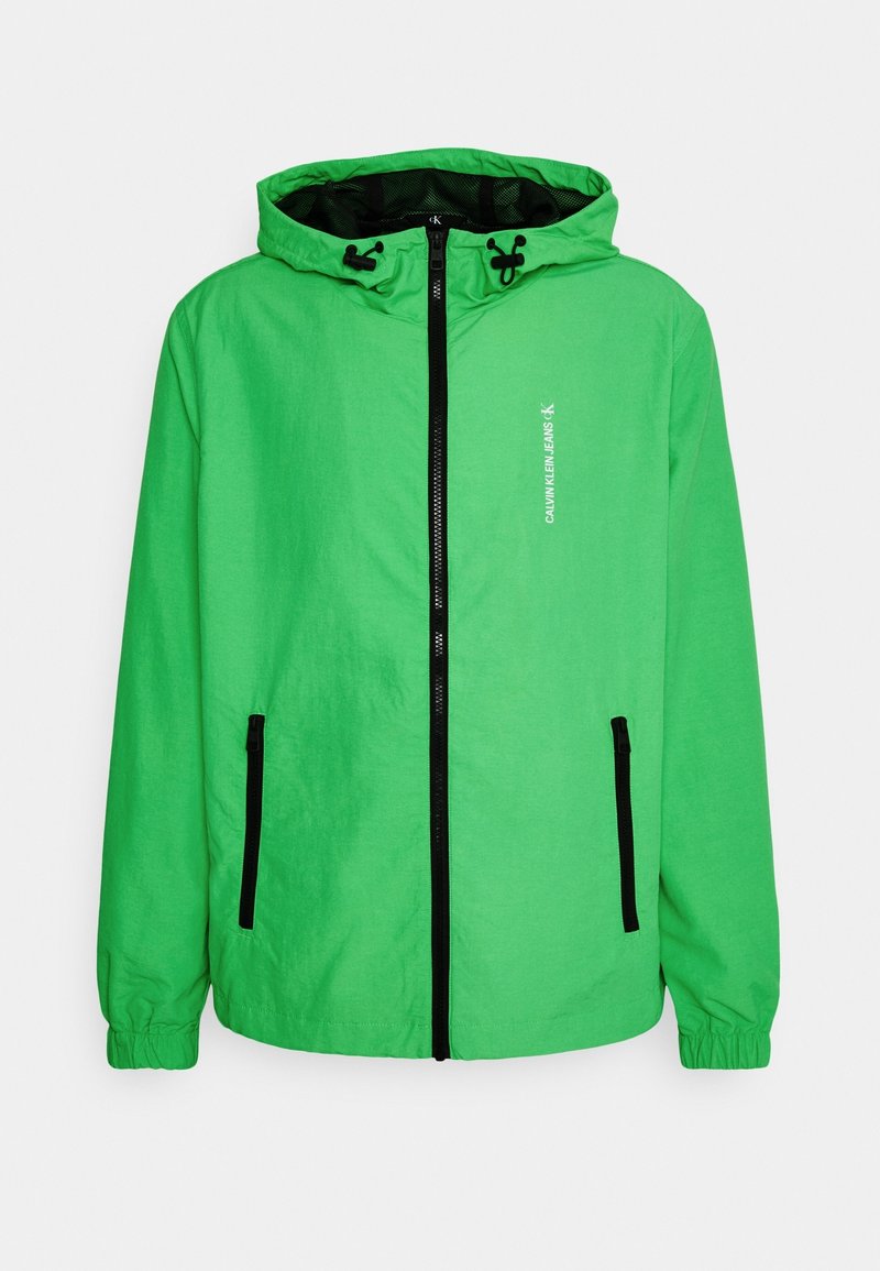 Groene capuchonjack gemaakt van lichtgewicht materiaal. Voorzien van een volledige rits, zwarte accenten en twee zijzakken.