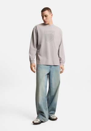 Młody mężczyzna w jasnoszarej koszulce z długim rękawem z logo, luźnych, wyblakłych niebieskich dżinsach i białych trampkach, stojący na jednolitym tle.