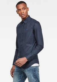 Camicetta blu navy con bottoni, dalla texture liscia, maniche lunghe e un piccolo logo azzurro sul petto. Abbinata a jeans blu sbiaditi.