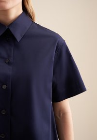 Granatowa koszula z krótkim rękawem, z kołnierzykiem zapinanym na guziki i przednim zapięciem. Gładki materiał z guzikami, o klasycznym, luźnym kroju.