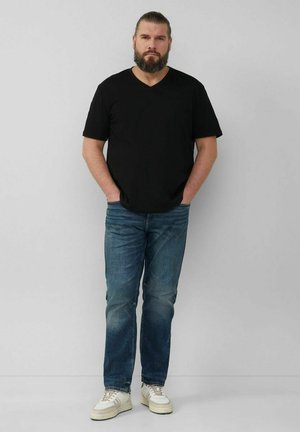 Black v-neck t-shirt paired with blue denim jeans. Model stands with hands in pockets, wearing white sneakers with tan accents.