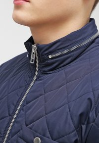 Zbliżenie na osobę w granatowej pikowanej kurtce z srebrnym suwakiem i zatrzaskiem na kieszeni piersiowej.