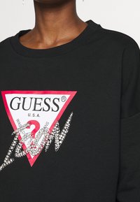 Felpa nera con scollo rotondo, caratterizzata da un grande logo a triangolo rosso con "GUESS" e "U.S.A." in bianco, decorato con rhinestones.