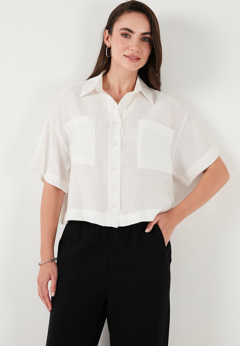 White short-sleeve, button-up shirt made of lightweight fabric with two front pockets and a collar, worn with black pants.