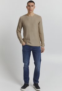 Beige strukturierter Pullover mit langen Ärmeln und rundem Halsausschnitt, kombiniert mit dunkelblauen Jeans und schwarzen Sneakers, vor einem neutralen Hintergrund stehend.