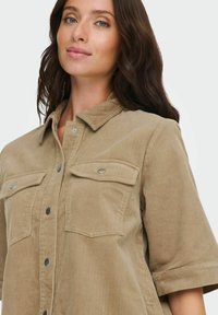 Svijetlo smeđa košulja jakna od korduroja s kratkim rukavima, džepovima na prsima i prednjim zatvaračem na kopče. Ima teksturirane pruge i srebrne metalne dijelove.