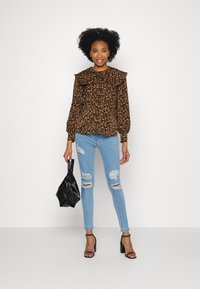 Blusa preta com padrão floral e decote franzido, mangas longas, combinada com jeans skinny claros rasgados e uma mala preta texturizada. Sapatos de salto pretos.