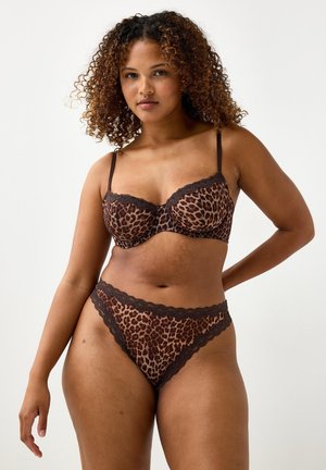 Kvinde med krøllet hår iført brunt og sort leopardmønstret lingeri med blonder, poserende med den ene hånd bag ryggen.