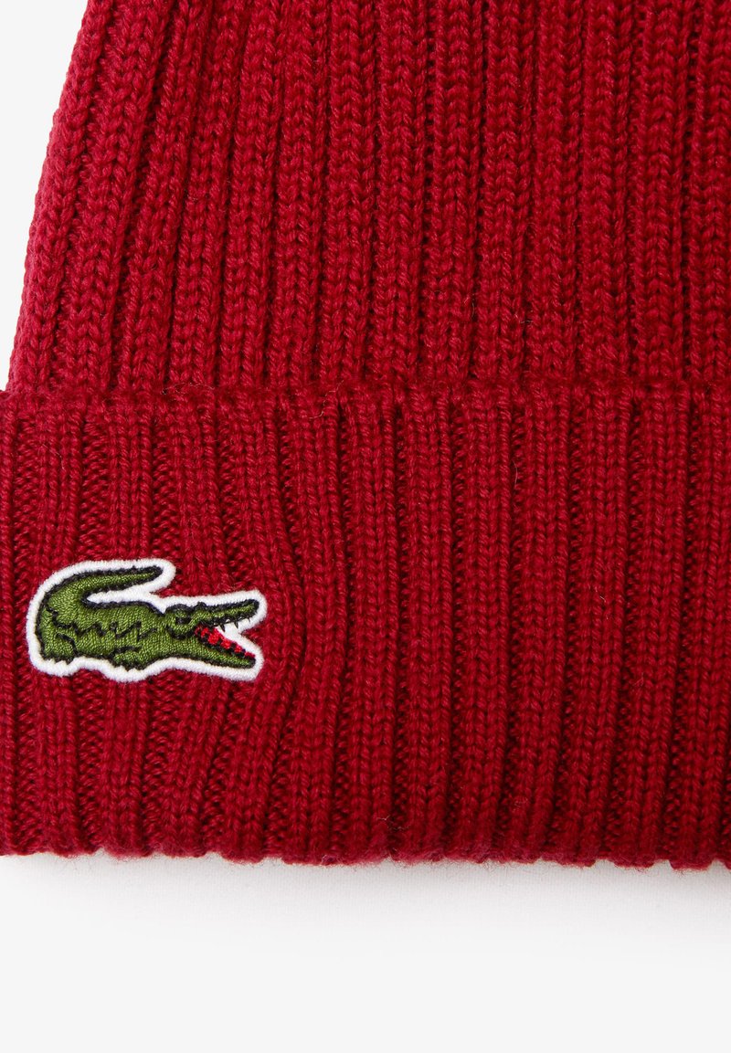 Rode gebreide beanie met een geribbeld patroon en een groen krokodillogo op een witte achtergrond.