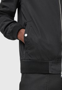 Czarna bomberka z elastycznymi mankietami, wyposażona w boczne kieszenie zabezpieczone białymi zatrzaskami. Gładka, lekka tkanina z dopasowanym dołem.