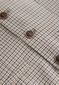 Tissu pied-de-poule marron et beige avec des boutons marron à quatre trous cousus, montrant une partie d'un vêtement sur mesure avec détail de poche.