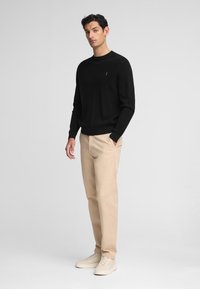 Suéter negro de punto con cuello redondo, chinos beige y zapatillas de color claro, de pie contra un fondo neutro.