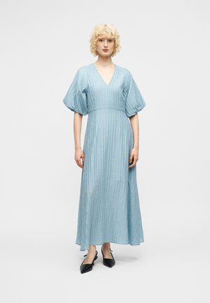 Kobieta o kręconych blond włosach ubrana w jasnoniebieską, teksturowaną sukienkę maxi z bufkami na rękawach oraz czarne, spiczaste buty typu slingback, stojąca na białym tle.