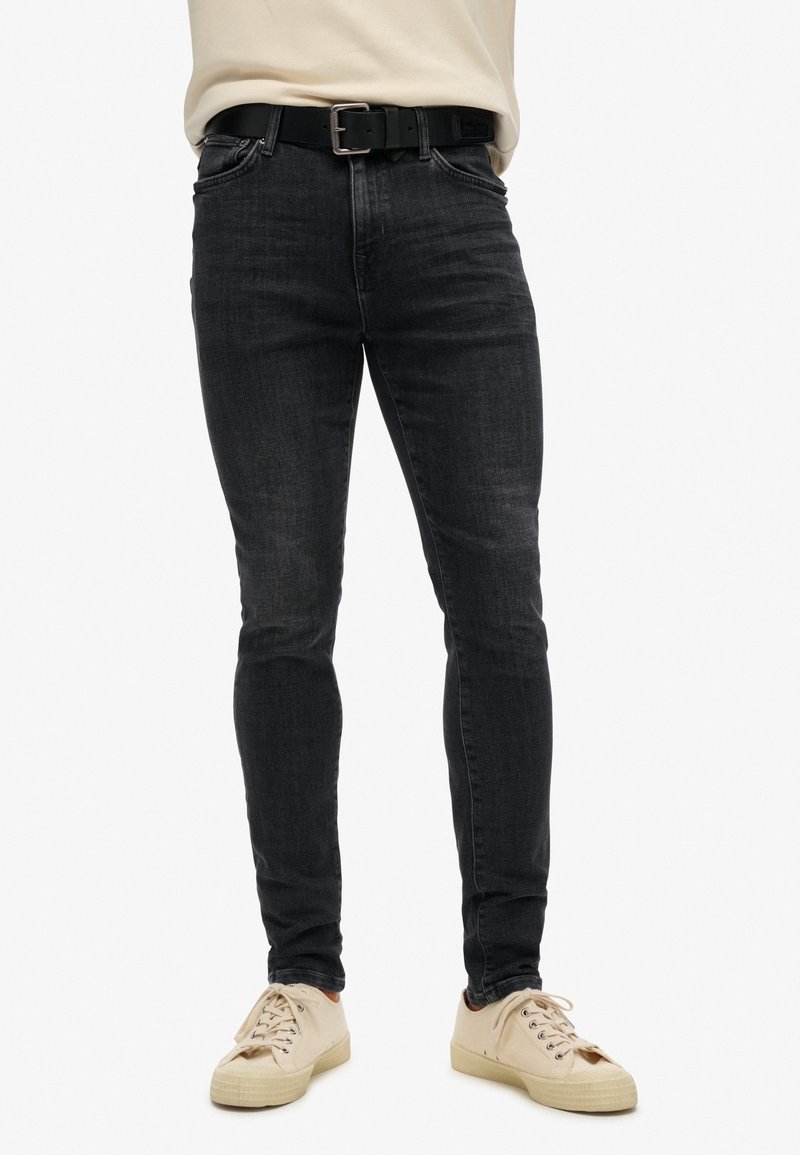 Jeans skinny negros de denim, con una cremallera estándar y un cinturón negro con hebilla plateada. Combinados con zapatillas beige.