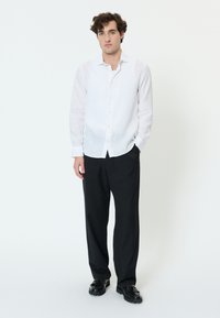 Mladenič stoji oblečen v belo srajco na gumbke, črne hlače in črne mokasine na enobarvnem svetlem ozadju.