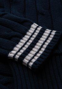 Maglione lavorato a maglia blu navy con un design a treccia testurizzata. Il polsino presenta righe orizzontali bianche e blu navy. Filato morbido e spesso.