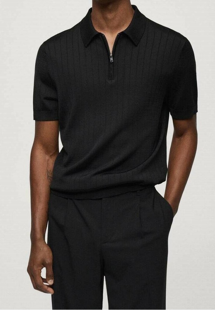 Homme portant un polo côtelé noir à manches courtes avec une fermeture éclair à l’avant et un pantalon noir, debout, une main dans la poche.