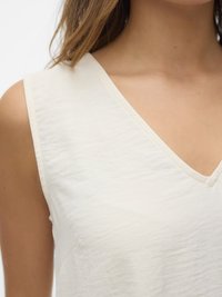 Frau trägt ein weißes ärmelloses Top mit V-Ausschnitt, das Schulter und oberen Brustbereich vor einfachem Hintergrund zeigt.