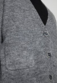 Detailní záběr na měkký šedý pletený cardigan s přední kapsou, černým vnitřním tričkem a třemi mramorově šedými knoflíky zapnutými.