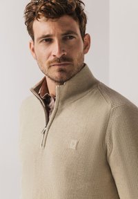 Beige gebreide trui met een halve ritssluiting, gestructureerde ribbelpatronen en een logopatch. Heeft een nauwsluitende pasvorm rondom de nek en schouders.