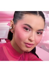 Junge Frau mit glattem dunklem Haar, trägt rosa Lidschatten, Rouge, Lippenstift, silberne Creolen und Perlenohrringe, gekleidet in einem rosa Oberteil vor rosa Wolken.