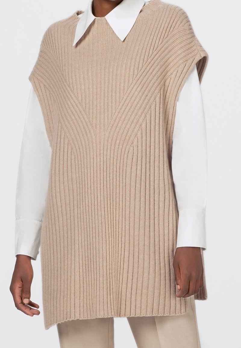 Beige gestrickte Sweaterweste mit einer gerippten Textur, V-förmigen Designdetails und kurzen Ärmeln, über einem weißen Hemd mit Kragen getragen.