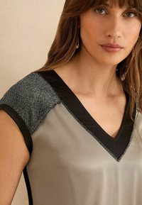 Kurzärmliges V-Ausschnitt-Oberteil aus einem gedämpften grauen Stoff mit schwarzem Besatz und strukturierten silbernen Akzenten an den Schultern. Glatte Textur im gesamten Shirt.