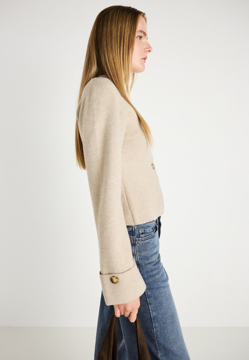 Beige, kort jakke med lange ermer og knappdetaljer, kombinert med blå jeans. Jevn tekstur og skreddersydd passform.