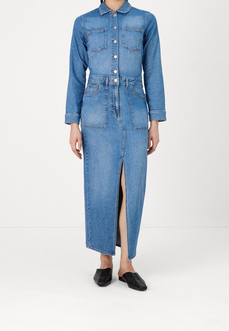 Femme portant une robe en denim bleu boutonnée avec poches devant et une fente avant, associée à des chaussures plates noires sans lacets.