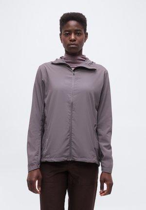 Νέο άτομο με κοντά μαλλιά που φοράει γκρι μπουφάν με φερμουάρ πάνω από ένα ανοιχτό μωβ πουλόβερ με λαιμό, στέκεται μπροστά σε ένα απλό λευκό φόντο.