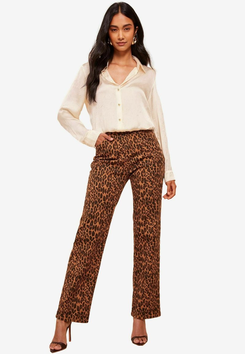 Cămașă deschisă la culoare, cu mâneci lungi, combinată cu pantaloni relaxați cu imprimeu de leopard maroniu și pantofi cu toc înalt, cu bretele.
