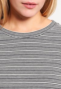 Tela a rayas en blanco y negro con textura acanalada, que presenta un amplio escote en collar. El patrón consiste en rayas de diferentes anchos.