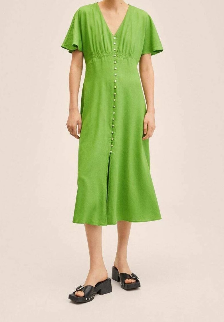 Femme portant une robe midi verte avec des manches courtes papillonnantes et des boutons sur le devant, associée à des sandales noires à talons ouverts avec des boucles.